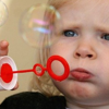 A young girl blowing bubbles, whilst a parent provides a guiding hand
